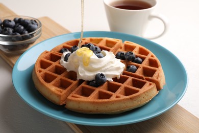 Pouring honey onto tasty Belgian waffle with blueberries and whipped cream on white table, closeup