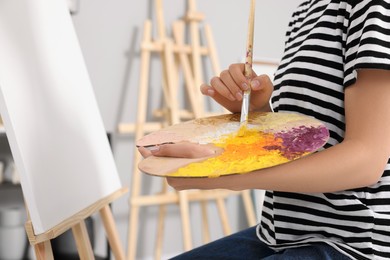 Photo of Woman mixing paints on palette with brush near easel in studio, closeup
