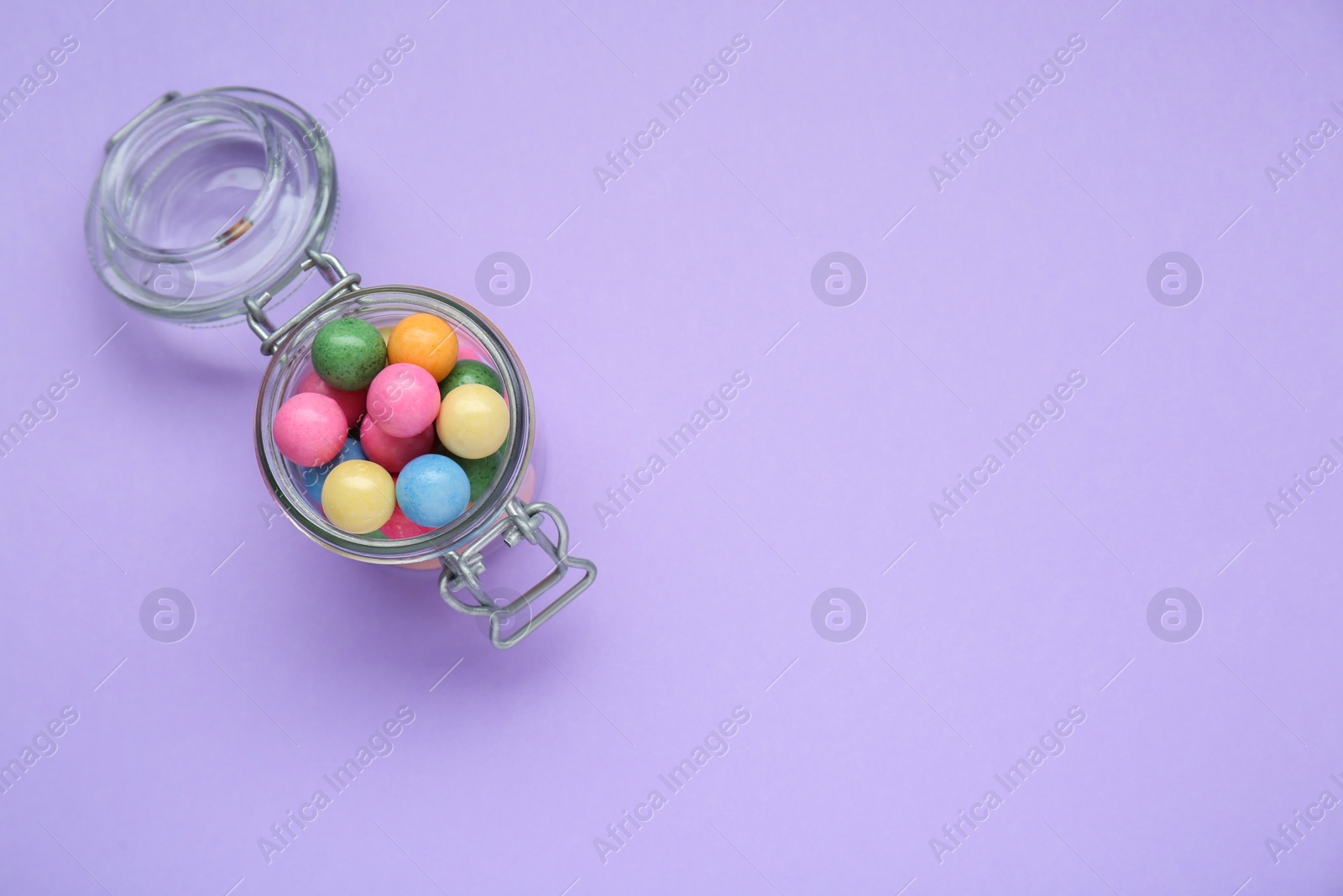 Photo of Jar with many bright gumballs on lilac background, top view. Space for text