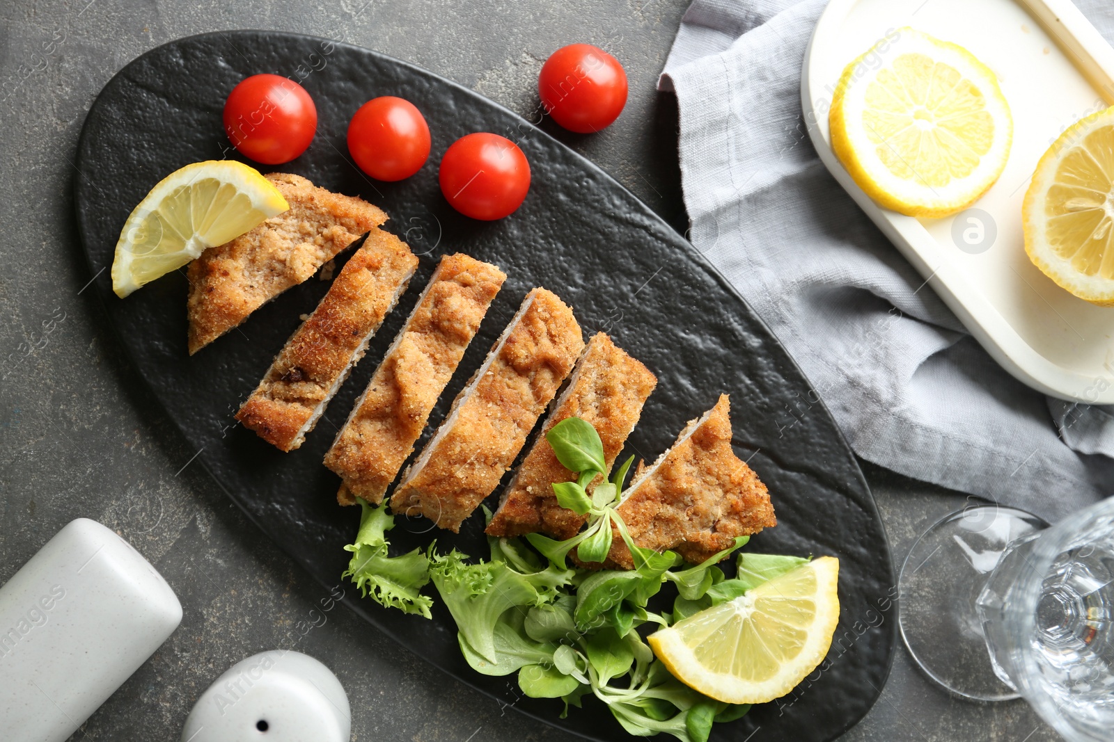 Photo of Tasty cut schnitzel served with tomatoes, greens and lemon on grey textured table, flat lay