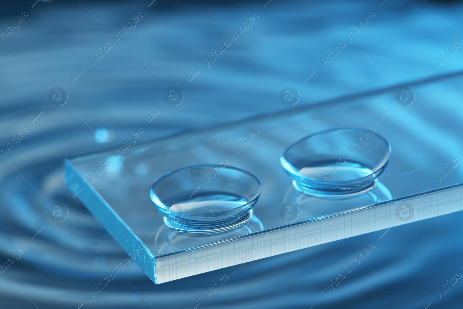 Photo of Pair of contact lenses on glass against blue background, closeup