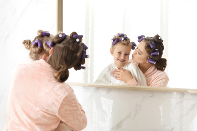 Photo of Happy mother and daughter with curlers near mirror in bathroom