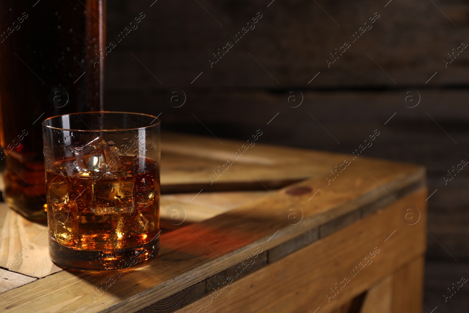 Photo of Whiskey with ice cubes in glass and bottle on wooden crate, closeup. Space for text