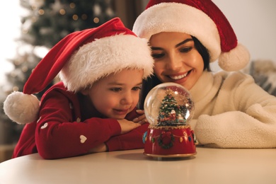 Mother and daughter in Santa hats playing with snow globe at table