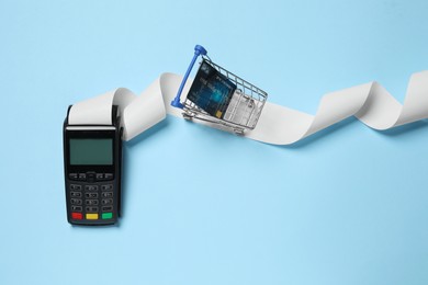 Photo of Payment terminal with thermal paper for receipt, small shopping cart and credit card on light blue background, flat lay