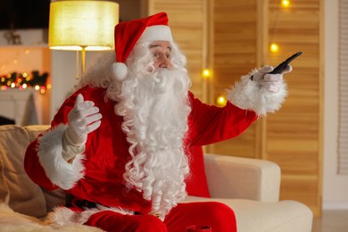 Merry Christmas. Santa Claus changing TV channels with remote control on sofa at home