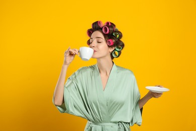 Beautiful young woman in silk bathrobe with hair curlers and cup of drink on yellow background