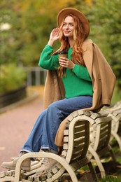 Photo of Beautiful woman drinking coffee from paper cup on bench in autumn park