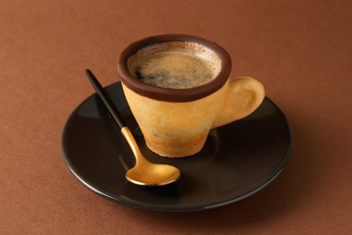 Photo of Delicious edible biscuit cup with coffee, spoon and saucer on brown background