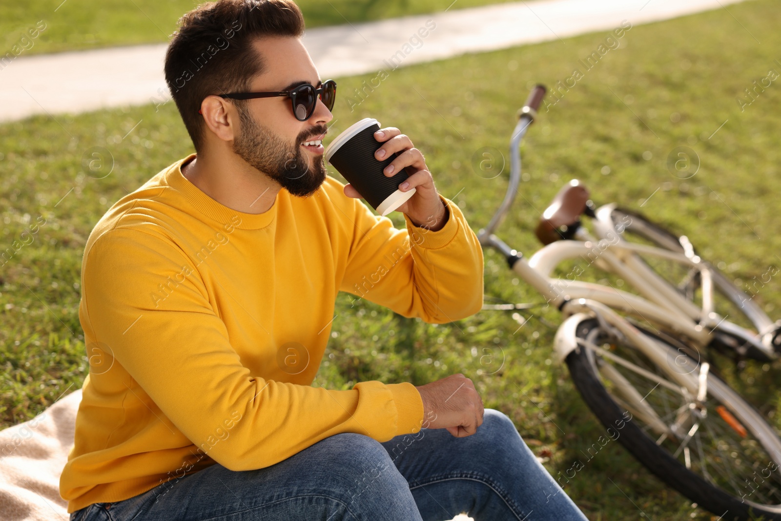 Photo of Young man sitting on green grass and drinking coffee near bicycle outdoors