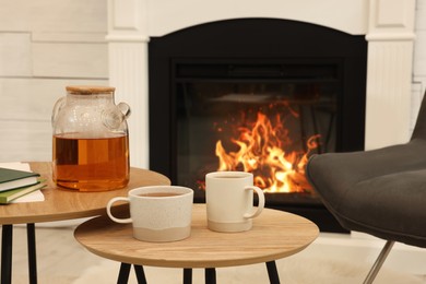 Cups of hot drink and teapot on wooden tables near decorative fireplace in room. Interior design