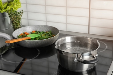 Pot near frying pan with vegetables on electric stove in kitchen