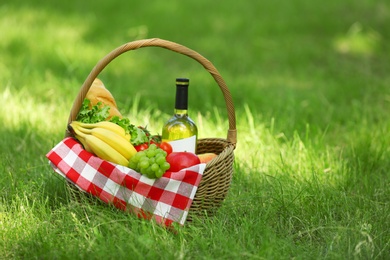 Wicker basket with blanket, wine and food on green grass in park, space for text. Summer picnic