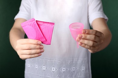 Woman holding menstrual cup and disposable pads on green background, closeup