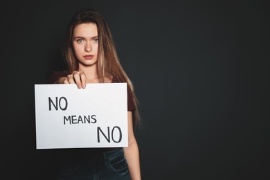 Photo of Young woman holding card with words NO MEANS NO against dark background. Space for text