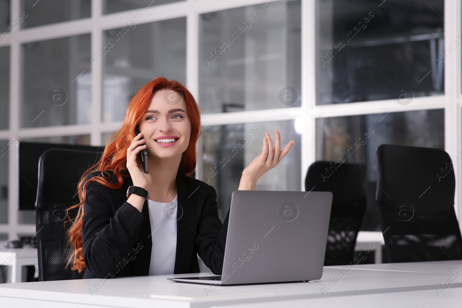 Photo of Happy woman talking on smartphone while working with laptop at white desk in office