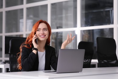 Photo of Happy woman talking on smartphone while working with laptop at white desk in office