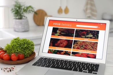 Photo of Modern laptop with open page for online food ordering on table in kitchen. Delivery service