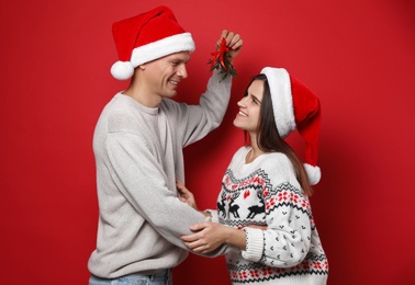 Photo of Happy couple in Santa hats standing under mistletoe bunch on red background