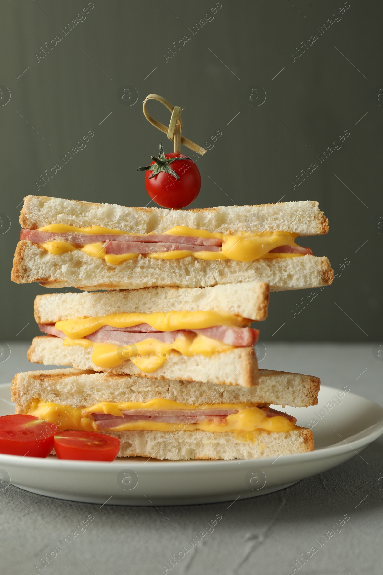 Photo of Stack of tasty sandwiches with ham and melted cheese served with tomatoes on grey textured table, closeup