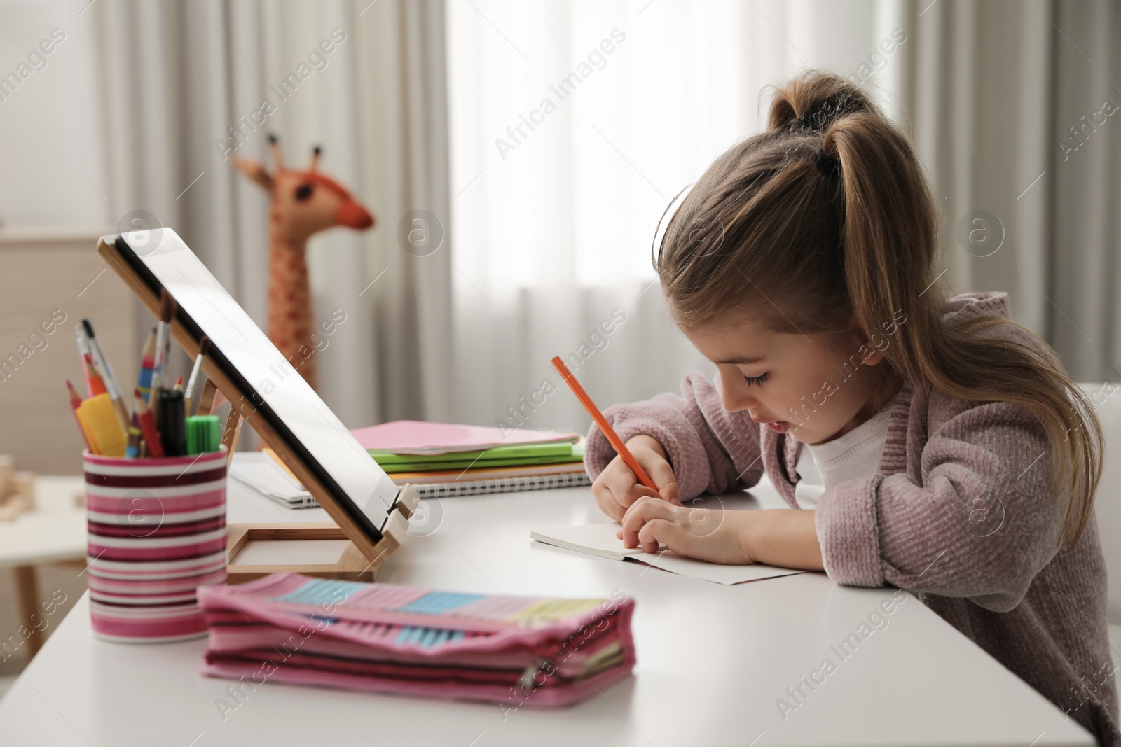 Photo of Adorable little girl doing homework with tablet at table indoors