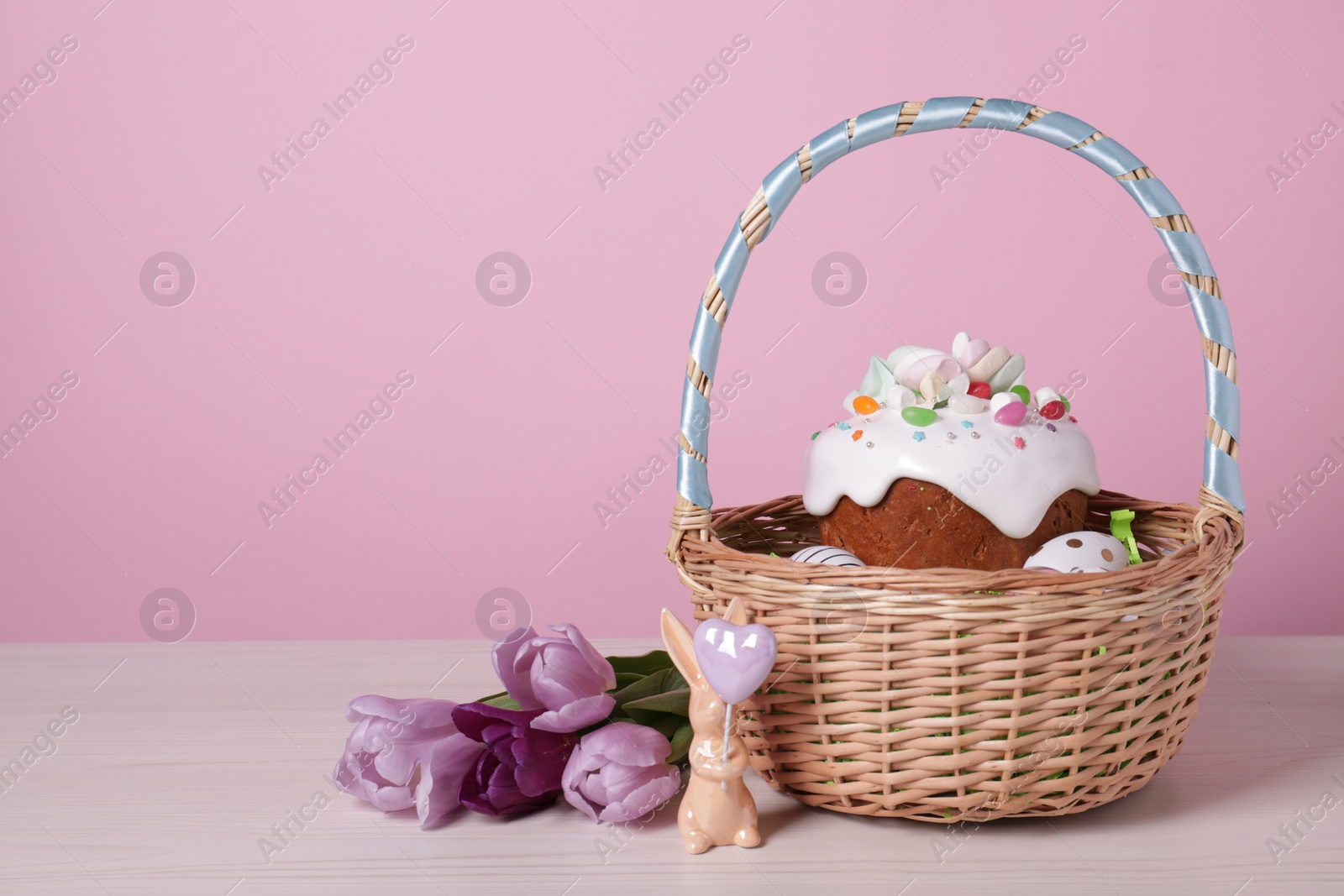 Photo of Easter basket with painted eggs, tasty cake, flowers and rabbit figure on white wooden table. Space for text