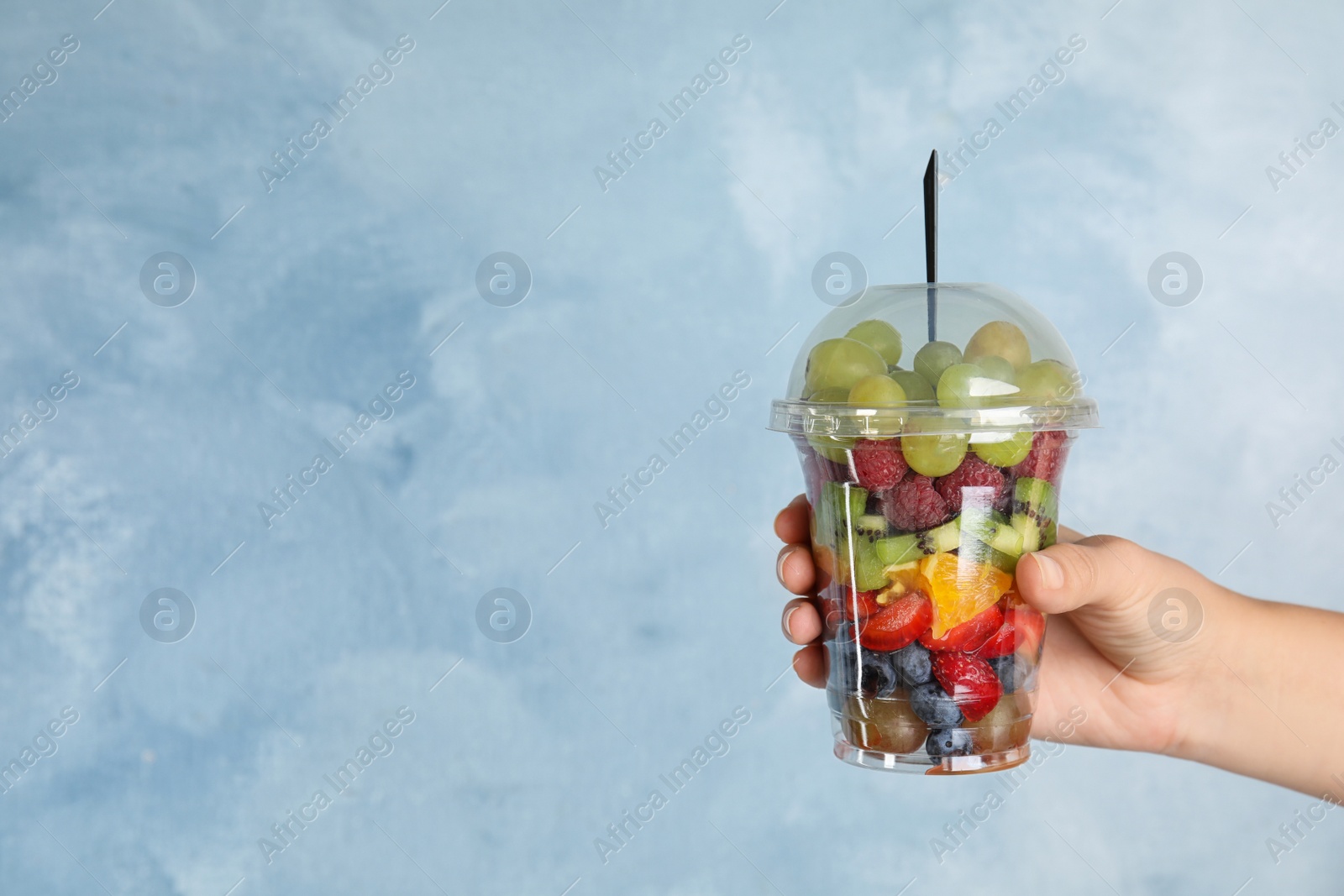 Photo of Woman holding plastic cup with fresh tasty fruit salad against light blue background, closeup. Space for text