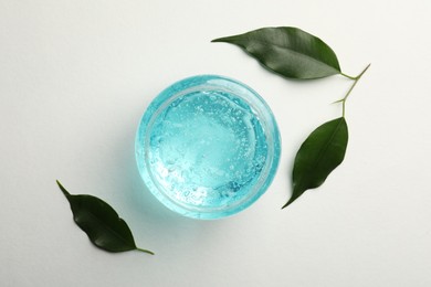 Jar of blue cosmetic gel and green leaves on white background, flat lay