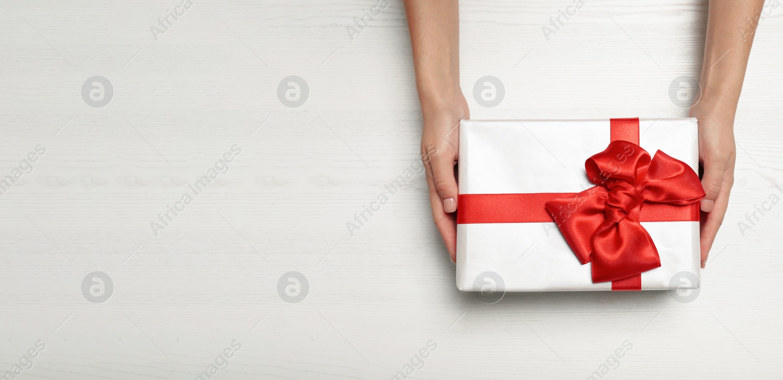 Image of Woman holding gift box with red bow and space for text on white wooden background, top view