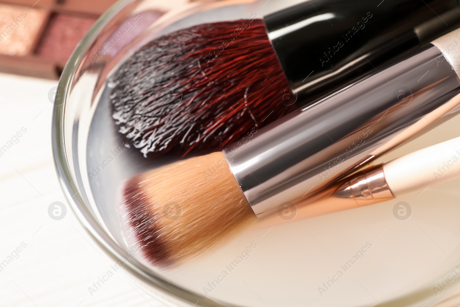 Photo of Cleaning makeup brushes in bowl with special liquid on white wooden table, closeup