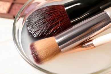 Photo of Cleaning makeup brushes in bowl with special liquid on white wooden table, closeup