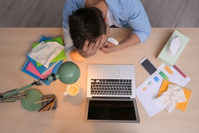 Photo of Exhausted man with tissue suffering from cold while working with laptop at table, top view