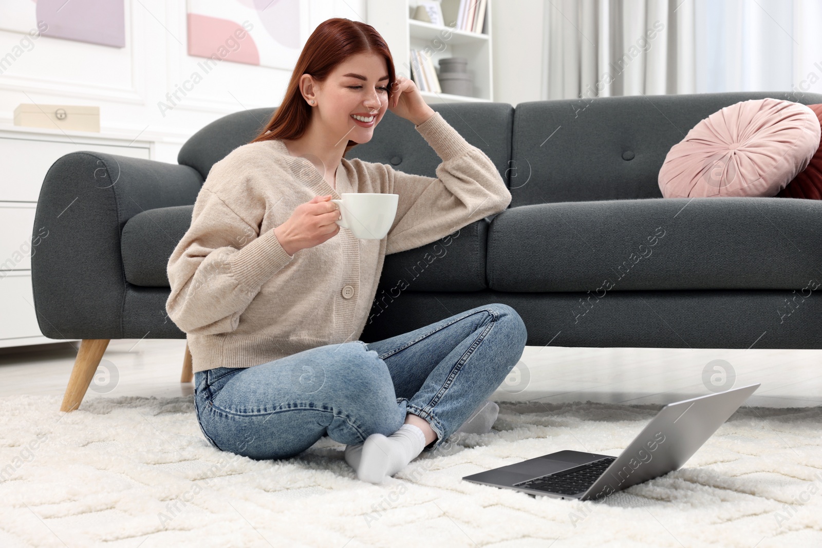 Photo of Happy woman with cup of drink and laptop on rug in living room