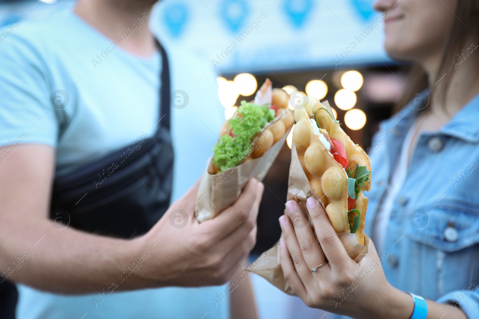 Photo of Young couple holding delicious bubble waffles with tomato and arugula outdoors, closeup