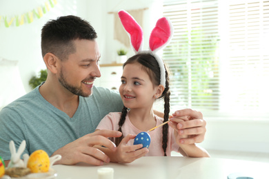 Happy daughter with bunny ears headband and her father painting Easter egg at home