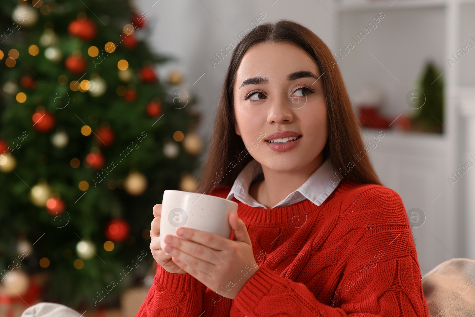 Photo of Woman holding cup of hot drink on sofa near Christmas tree indoors