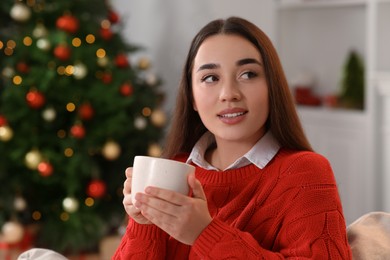 Woman holding cup of hot drink on sofa near Christmas tree indoors