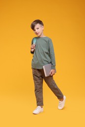 Photo of Cute schoolboy with book walking on orange background