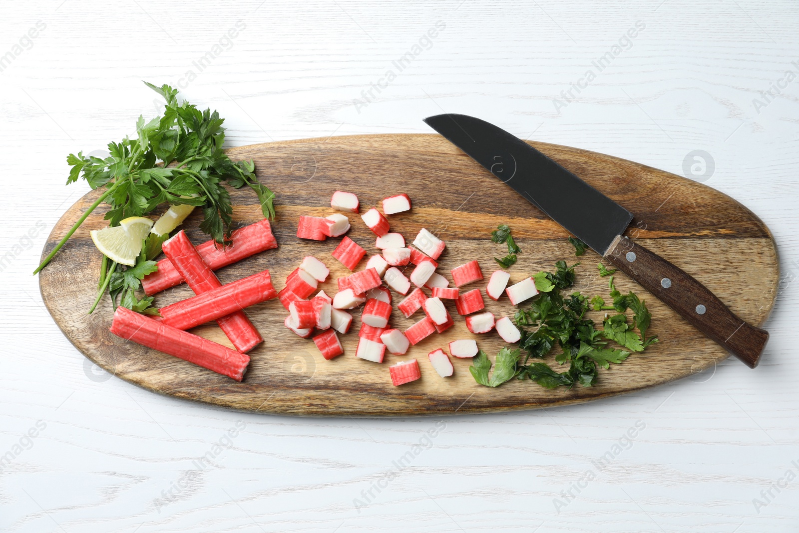 Photo of Delicious crab sticks with parsley, lemon and knife on white wooden table, top view