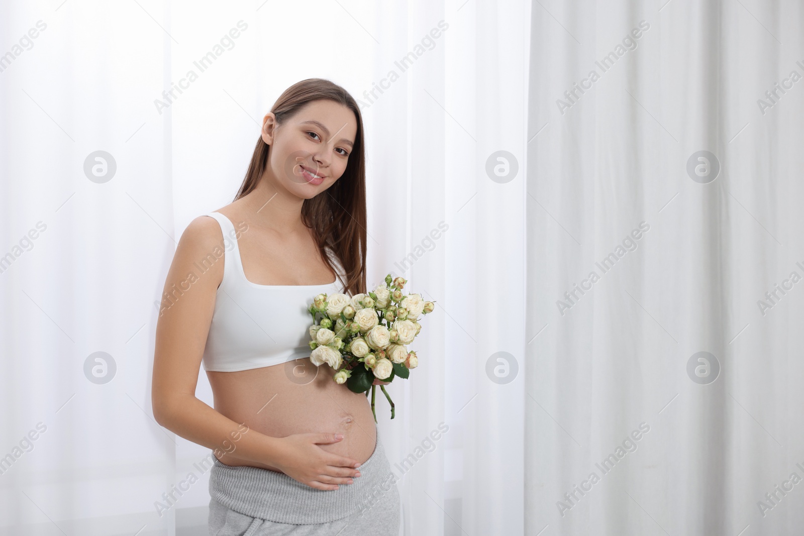 Photo of Beautiful pregnant woman with bouquet of roses near window indoors, space for text