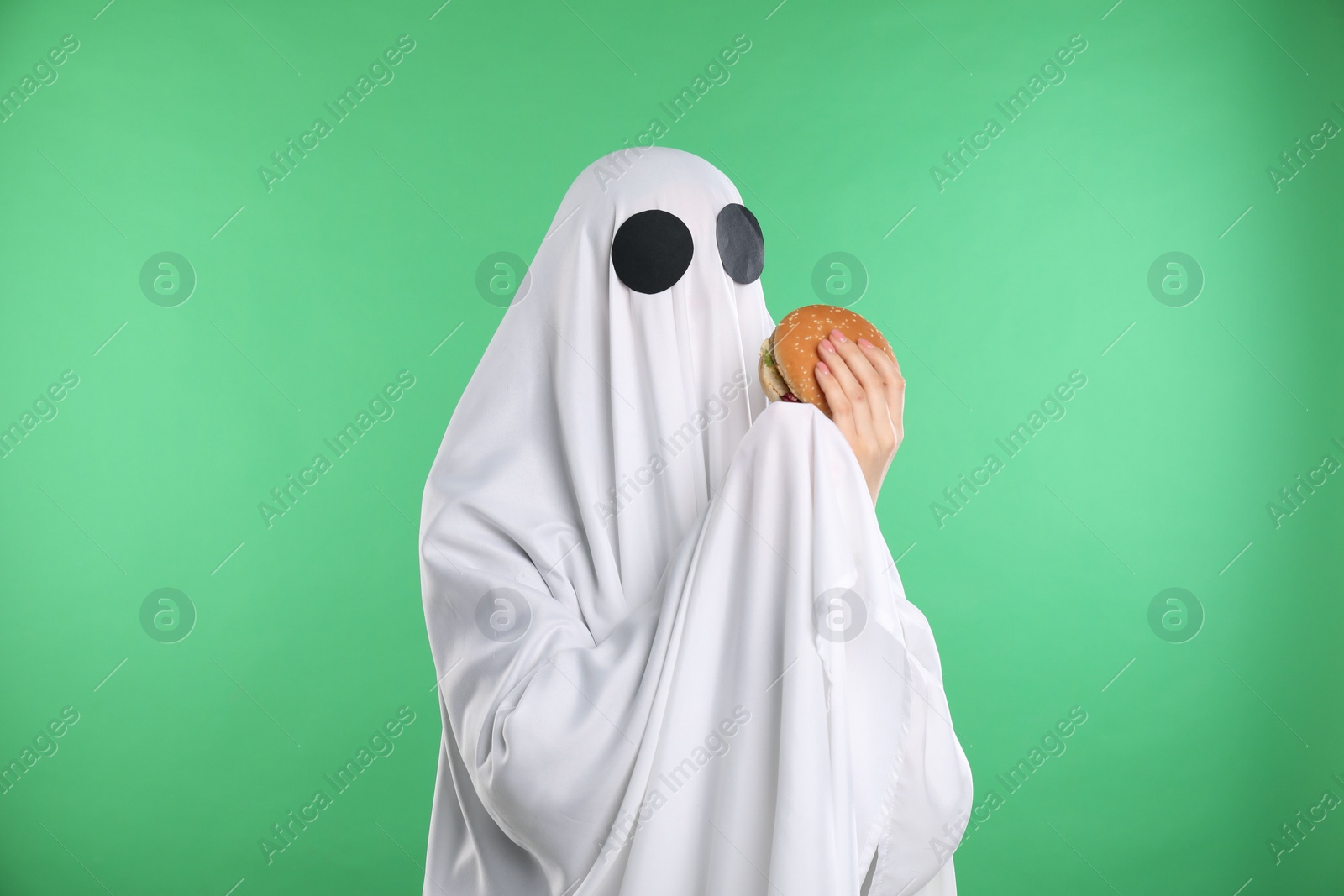 Photo of Creepy ghost. Person in white sheet with tasty hamburger on green background