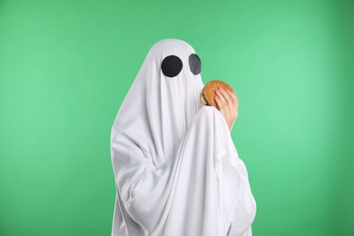 Photo of Creepy ghost. Person in white sheet with tasty hamburger on green background