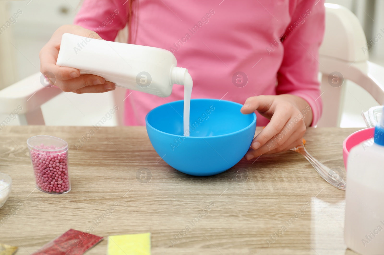 Photo of Little girl pouring glue into bowl at table in room, closeup. DIY slime toy