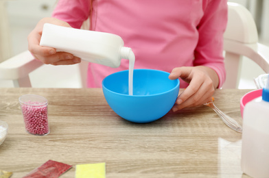 Photo of Little girl pouring glue into bowl at table in room, closeup. DIY slime toy