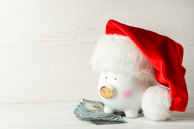 Photo of Piggy bank with Santa hat and dollar banknotes on white wooden table. Space for text