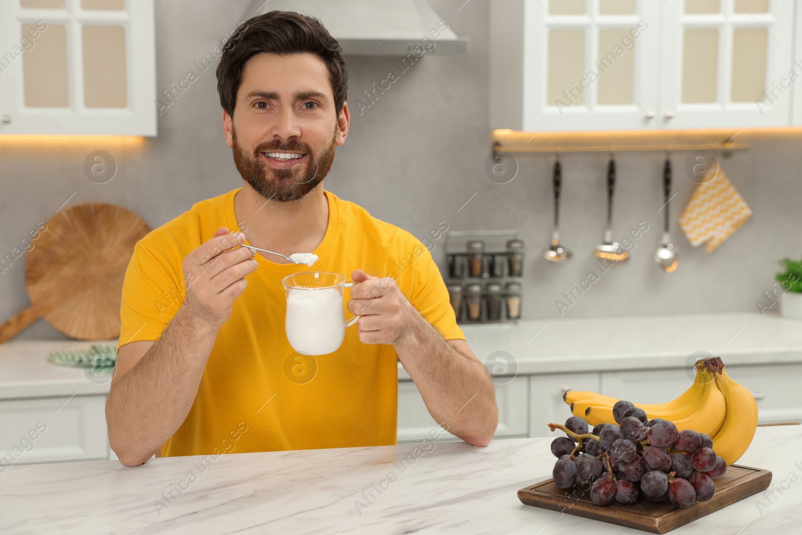 Photo of Handsome man with delicious yogurt at white marble table in kitchen. Space for text