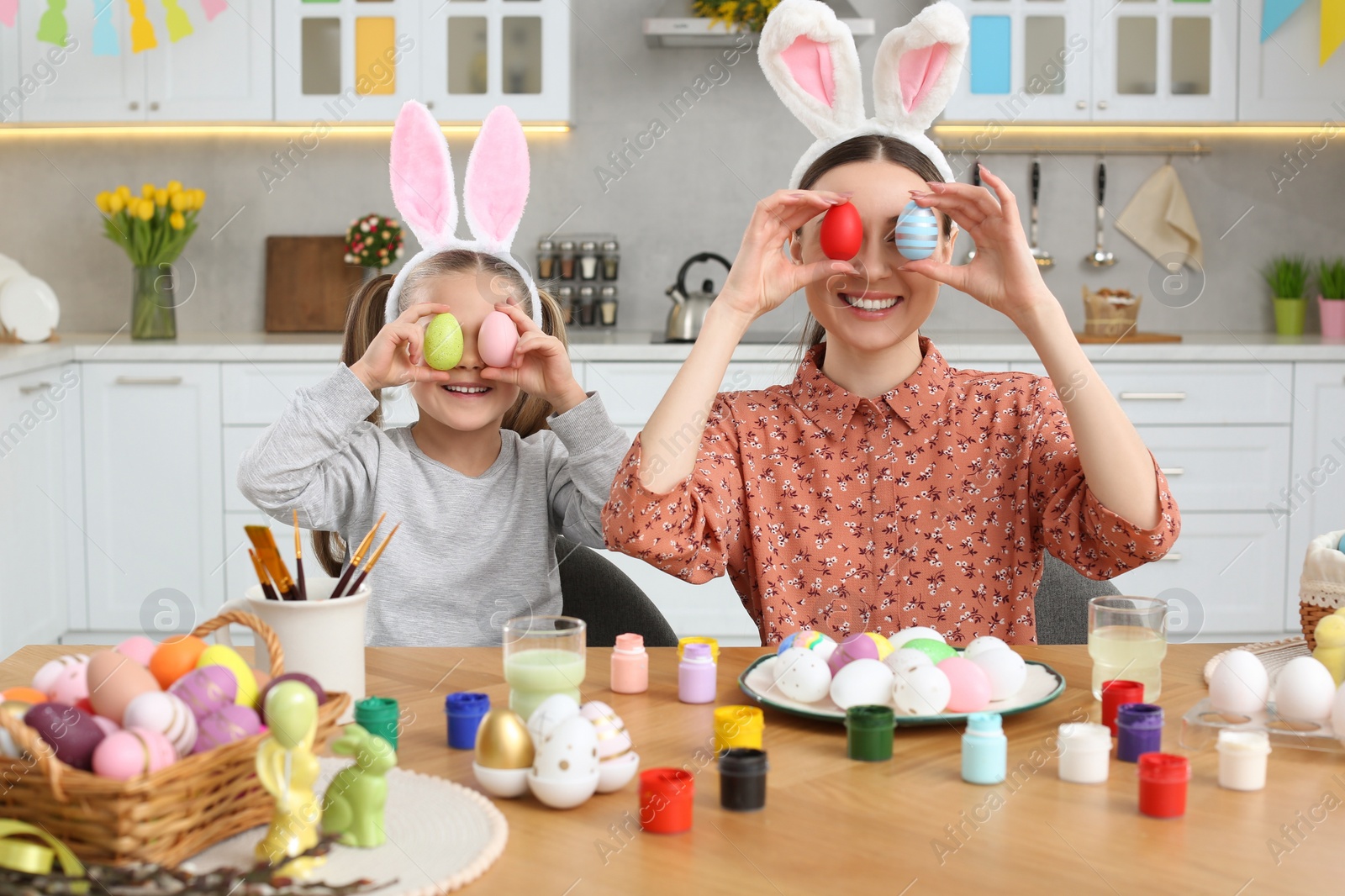 Photo of Mother and her cute daughter covering eyes with beautifully painted Easter eggs at table in kitchen