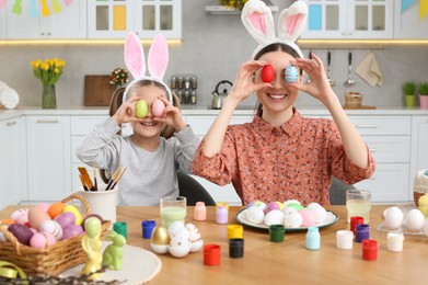 Mother and her cute daughter covering eyes with beautifully painted Easter eggs at table in kitchen