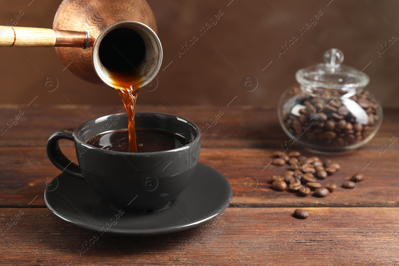 Photo of Turkish coffee. Pouring brewed beverage from cezve into cup at wooden table against brown background