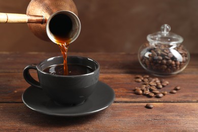 Photo of Turkish coffee. Pouring brewed beverage from cezve into cup at wooden table against brown background